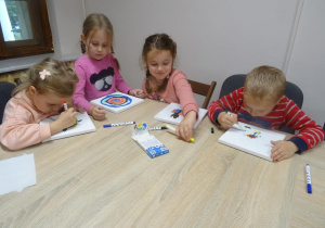 Czworo dzieci siedzi przy stole. Dwoje z nich trzyma mazaki w ręku i kończy malowanie płótna. Jedna dziewczynka odkłada mazak na stół a druga spogląda na swoją pracę.